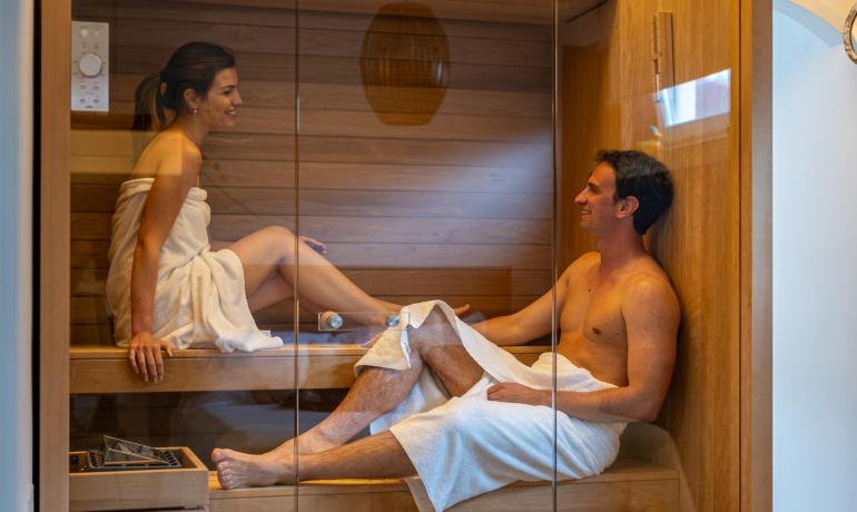 Coppia felice nella sauna, avvolti in asciugamani, sorrisi e relax.